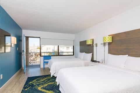 Hotel bedroom with two queen beds, teal accent wall, wooden headboards, and balcony with city view