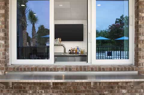 Indoor-outdoor bar setup with wall-mounted TV, beverage station, and palm tree views through glass doors