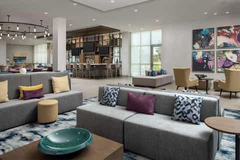 Modern hotel lobby with gray sectional sofas, colorful artwork, bar area, and large windows overlooking grounds
