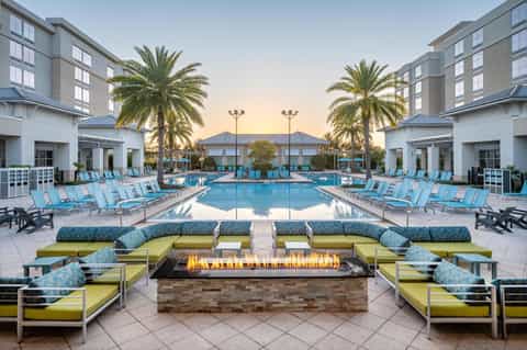 Resort pool at sunset with turquoise water, lounge chairs, firepit, and palm trees flanking modern buildings