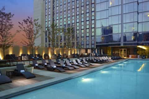 Rooftop pool at dusk with lounge chairs, illuminated high-rise hotel building, and city views