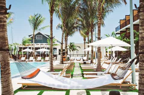 Tropical resort pool area with palm trees, hammocks, white umbrellas, and lounging chairs around turquoise water