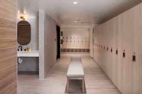 Modern spa locker room with wooden lockers, vanity station with oval mirror, and bench seating along a light wood corridor