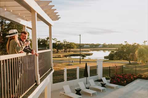 Couple on pergola balcony toasting with wine overlooking marshland and water with trees