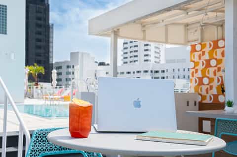 Rooftop workspace with laptop, cocktail, and orange fabric decor overlooking city skyline and pool