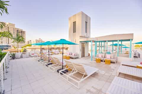 Rooftop pool deck with turquoise umbrellas, white loungers, and coastal city views at sunset