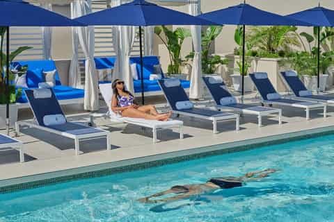 Resort pool area with lounge chairs, blue umbrellas, white cabanas, and swimmers in crystal-clear water