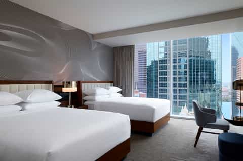 Modern hotel room with two queen beds, city skyline views through floor-to-ceiling windows, and abstract wall art