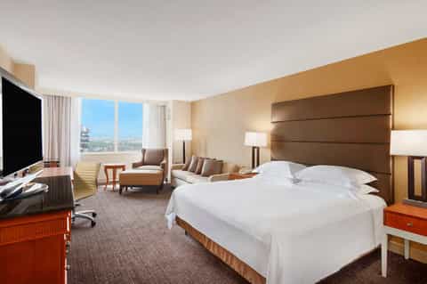 Upscale hotel bedroom with king bed, brown headboard, sitting area, desk, and panoramic city views through large windows