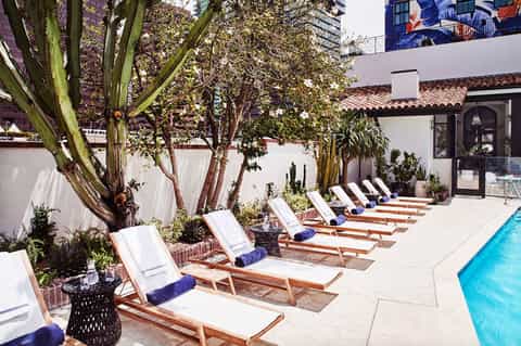 Poolside lounge area with wooden deck chairs, white cushions, and large trees providing shade