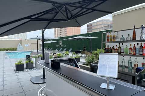 Rooftop bar with black umbrella, bottle display, and pool area with green hedge wall backdrop