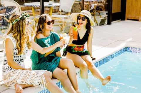 Three women in swimwear enjoying drinks poolside at a luxury resort with white lounge chairs