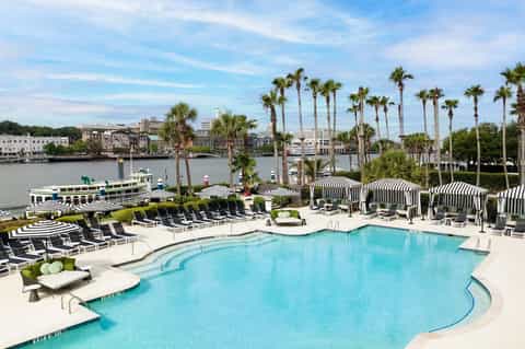 Resort pool with blue water, striped umbrellas, palm trees, and riverfront views with boats and city skyline