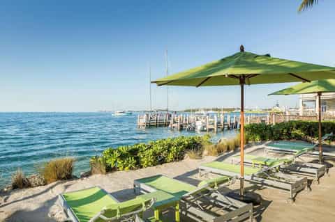 Beachfront lounge area with green umbrellas, lounge chairs, sailboats, and marina dock overlooking calm blue water