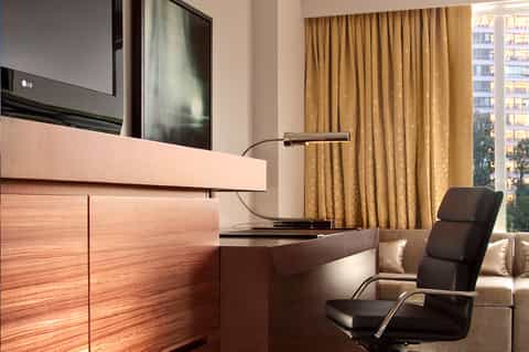 Modern hotel room desk with wood paneling, TV, black chair, gold lamp, and city view through curtains