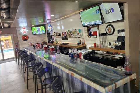 Sports bar interior with long counter, bar seating, TV screens, and shelving with bottles and sports memorabilia
