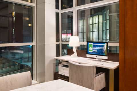 Hotel room desk with computer monitor and lamp, overlooking city skyline through floor-to-ceiling windows