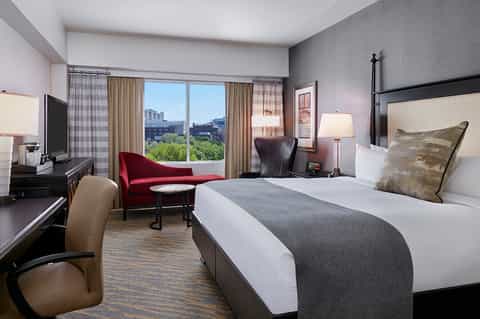 Modern hotel bedroom with king bed, city views from large window, burgundy accent chair, and contemporary furnishings