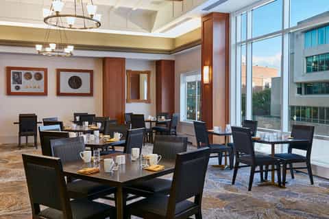 Modern breakfast room with black chairs, wooden tables, decorative wall clocks, and floor-to-ceiling windows with city views