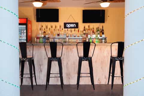 Modern bar interior with yellow walls, black bar stools, and shelves of liquor bottles beneath mounted TVs