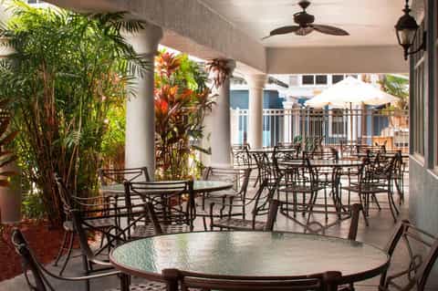 Covered outdoor patio with round tables, metal chairs, tropical plants, ceiling fans, and white umbrellas visible beyond
