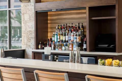 Modern hotel bar with extensive liquor selection, wooden backing, white counter, and bar seating