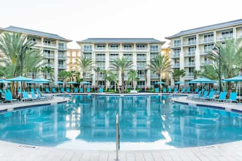 Large resort pool with bright turquoise water, palm trees, colorful lounge chairs, and multi-story hotel buildings