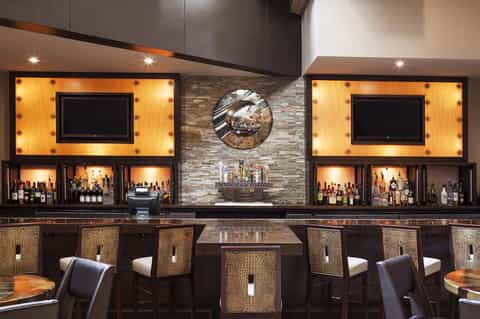 Sophisticated hotel bar with illuminated shelving, TVs, stone accent wall, and woven bar stools