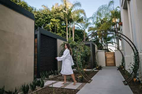 Woman in white robe walking through modern spa courtyard with black louvered gates