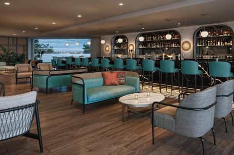 Modern hotel bar and lounge with teal seating, pendant lighting, and floor-to-ceiling windows overlooking water