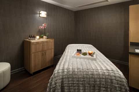 Spa treatment room with massage table, wooden furniture, flowers, and dark textured walls
