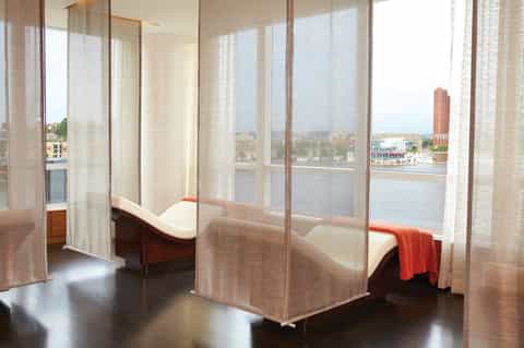 Spa treatment room with floor-to-ceiling windows overlooking waterfront, sheer curtains, and two massage beds