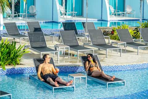 Two guests relaxing on lounge chairs in shallow pool with tropical setting and blue umbrellas