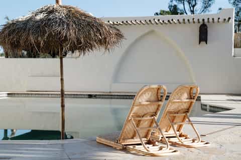 Minimalist poolside lounge with two woven chairs under thatched umbrella near white building