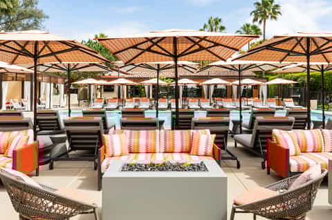 Poolside lounge area with fire pit, orange and striped cushions, large umbrellas, and palm trees