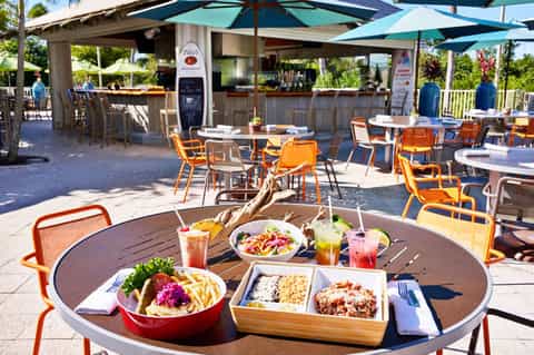 Outdoor beachfront restaurant with orange chairs, colorful dishes, tropical flowers, and ocean view