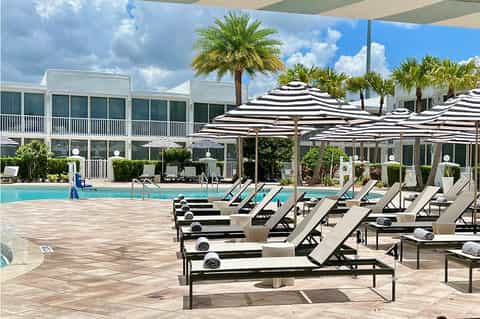 Resort pool deck with striped umbrellas, lounge chairs, palm trees, and white building