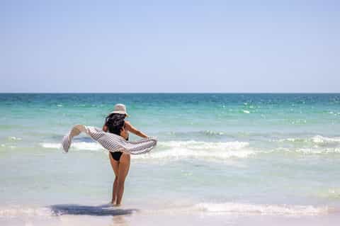 Woman in straw hat wading in turquoise ocean waves on sandy beach