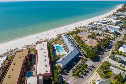 Aerial view of beachfront resort with pool, sandy beach, palm trees, and surrounding accommodations