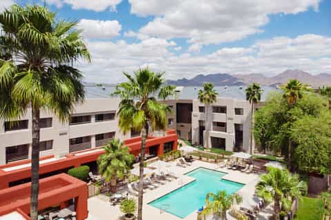 Modern resort courtyard with turquoise pool, palm trees, white multi-story buildings, and mountain views in the background
