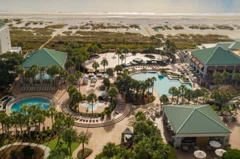 Aerial view of beachfront resort with multiple pools, palm trees, green roofs, and sandy beach