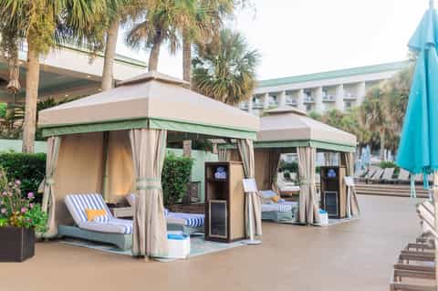 Beachside cabana lounge with green and cream canopies, lounge chairs, palm trees, and hotel building in background