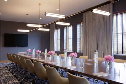Modern conference room with long table, tan chairs, pink flowers, and flat-screen TV