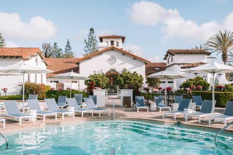 Outdoor pool area with white umbrellas, lounge chairs, and Spanish-style resort buildings