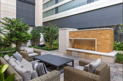 Modern outdoor courtyard with fireplace, woven lounge chairs, dark dining table, and greenery