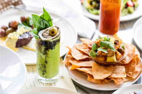 Fine dining table with green cocktail garnished with oyster, plated seafood dishes, and tropical drink
