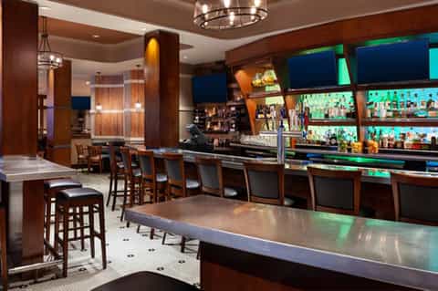Modern hotel bar and lounge with dark wood finishes, multiple TV screens, illuminated shelving, and bar seating