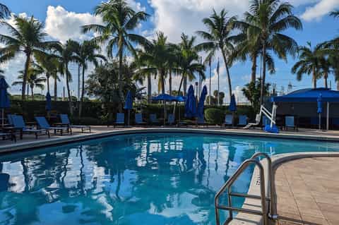 Resort pool with blue umbrellas and lounge chairs surrounded by palm trees and clear sky