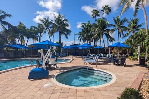 Resort pool with hot tub, blue umbrellas, palm trees, loungers, and desert landscape in background