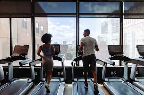 Two people running on treadmills in high-rise gym with floor-to-ceiling windows overlooking city skyline